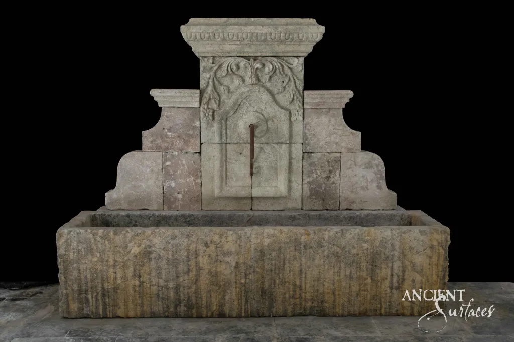 Wide grey limestone wall fountain with a moulded cornice crest over a long stone trough
